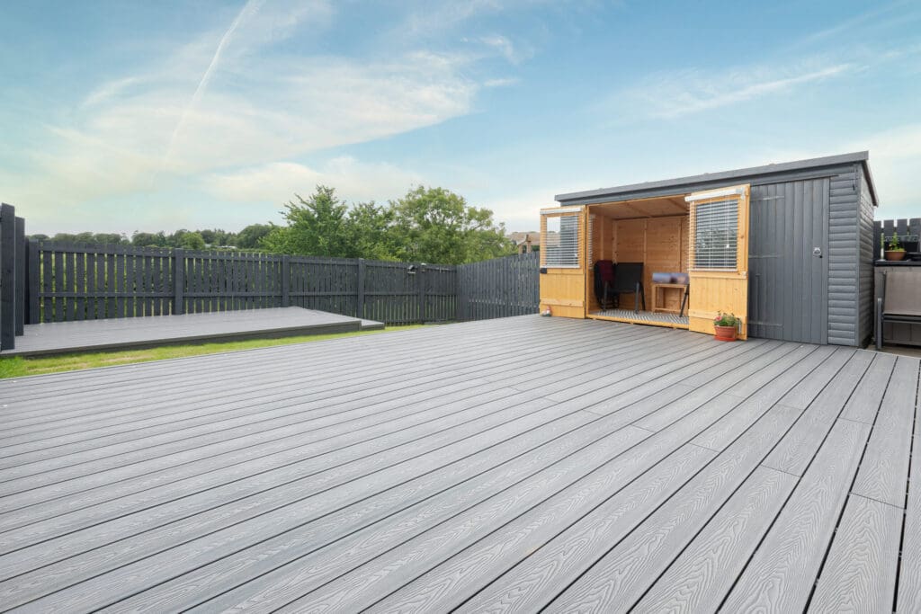 A spacious outdoor deck with gray wooden flooring, showcasing expert decking craftsmanship. It features a small open shed with wooden and gray walls. The area is bordered by a dark fence and complemented by a green lawn with trees in the background under a blue sky.