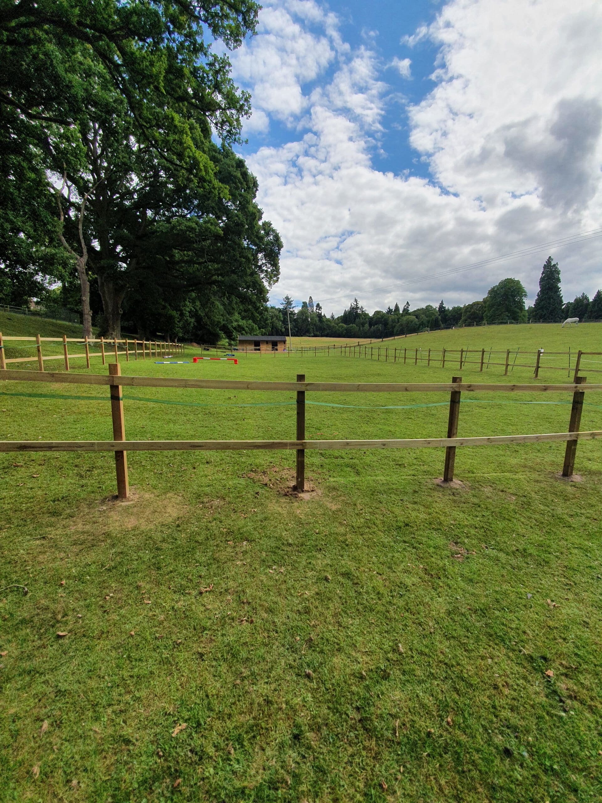 A wide, grassy field bordered by a wooden fence under a partly cloudy sky. Trees stand in the background, while red markers dot the lawn like a landscaper's thoughtful touch, hinting at future plans for driveways or decking.