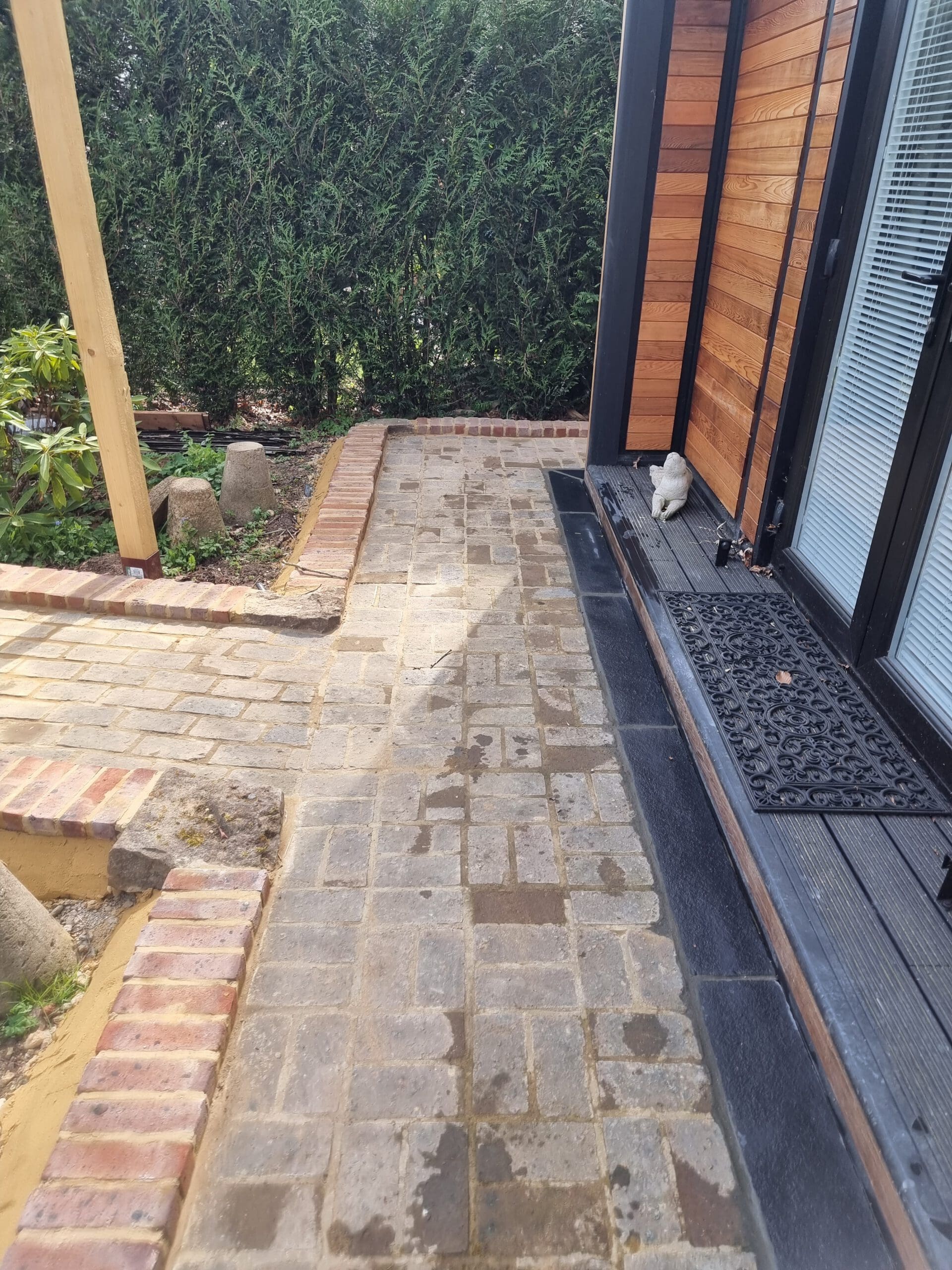 A brick pathway next to a modern wooden building with black trim, crafted by skilled landscapers, is bordered by short brick walls and lush greenery. A white object, possibly a statue, sits on a black mat by the building's entrance.