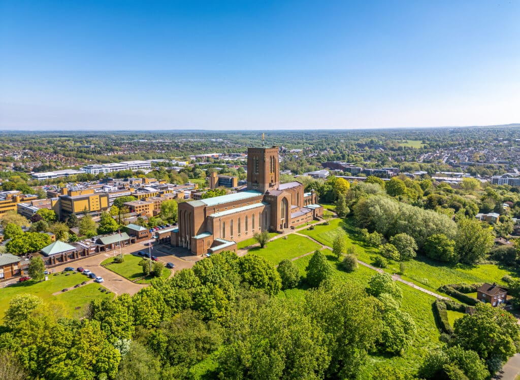 Aerial view of a large brick cathedral surrounded by lush, green gardens and trees, crafted by a skilled landscaper. In the background, new fencing borders the sprawling town under a clear, blue sky. The scene is bright and tranquil, highlighting the cathedral's architecture and the town's layout.