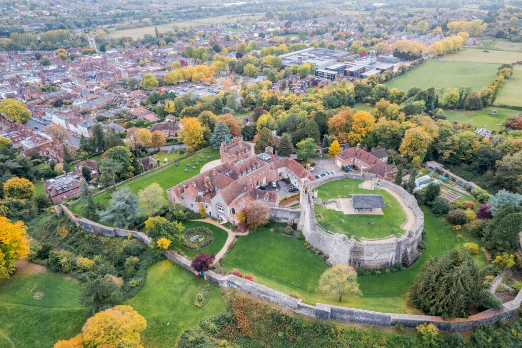 Aerial view of a historic castle with circular stone walls, complemented by surrounding greenery and new fencing. The area features trees in stunning fall colors, transitioning into a residential neighborhood. A landscaper's touch extends to a small town with roads and houses in the background.