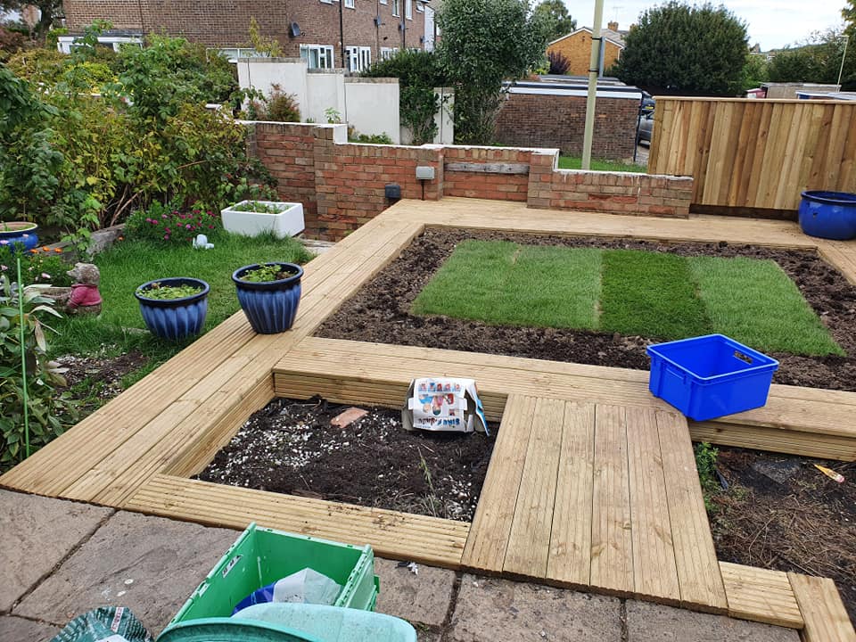 A garden under renovation with newly laid turf, wooden decking paths, blue planters, and plastic tubs. Tools and containers are scattered around, and a brick wall separates the garden from the street.