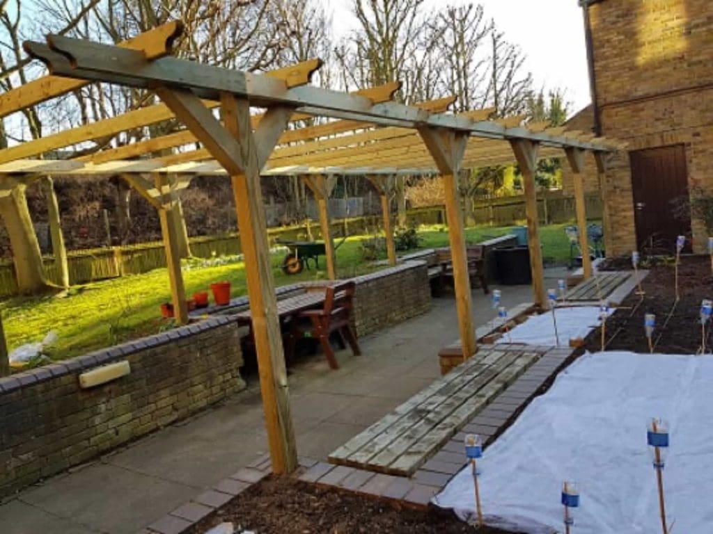 Wooden pergola frame under construction in a backyard; raised brick garden beds, patio furniture, garden tools, and covered planting beds are visible, with trees and a lawn in the background.