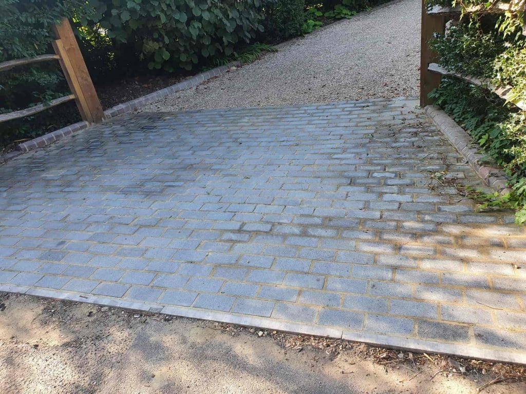 A paved path made of rectangular gray bricks leads to a gravel pathway, bordered by wooden railings and green bushes on either side. Sunlight casts shadows across the brick surface.