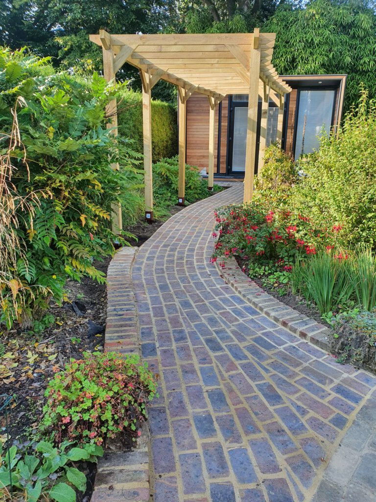 Curved brick pathway through a lush garden with green plants and red flowers, leading to a modern wooden garden building under a light wooden pergola.