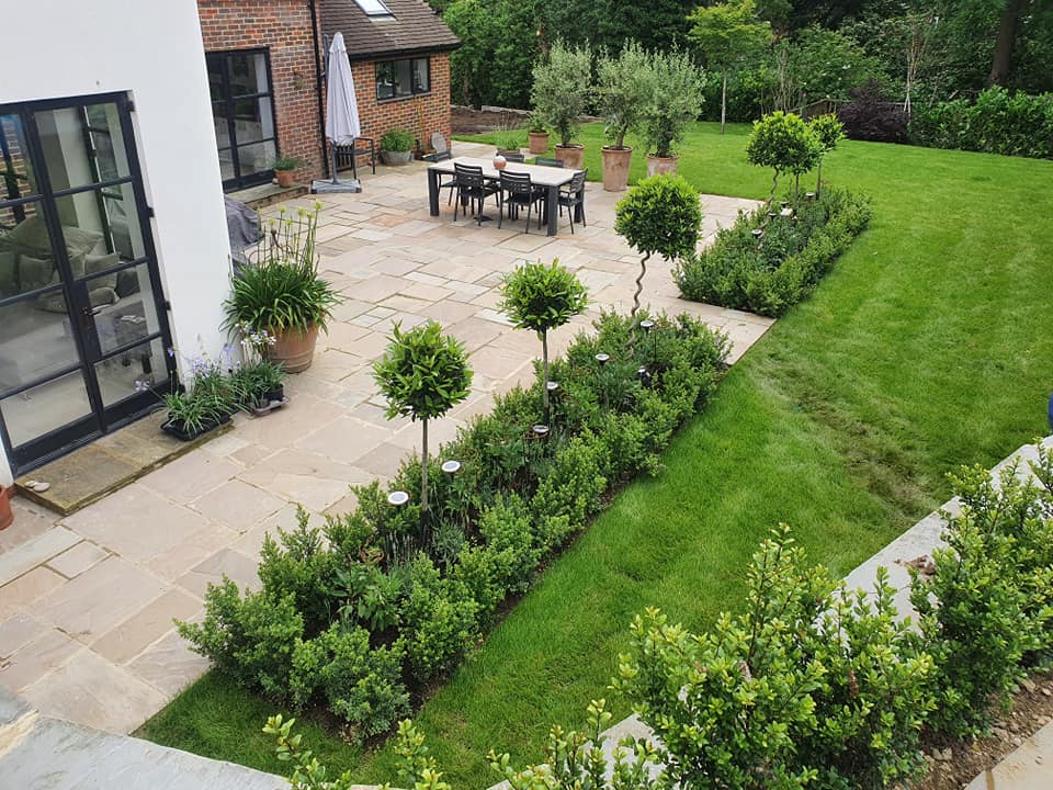 A modern patio with stone tiles, outdoor dining table, potted plants, trimmed hedges, small trees, and a manicured lawn next to a brick and white house surrounded by lush greenery.