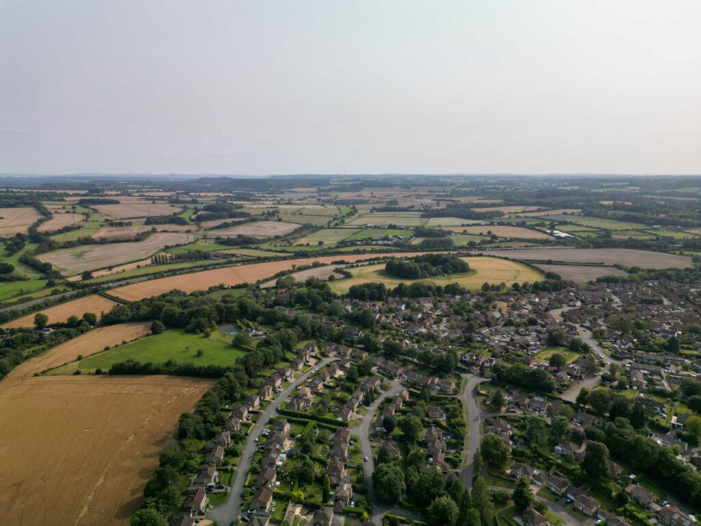 Aerial view of a rural landscape featuring a village with houses, winding roads, green trees, and surrounding fields stretching into the distance under a hazy sky.