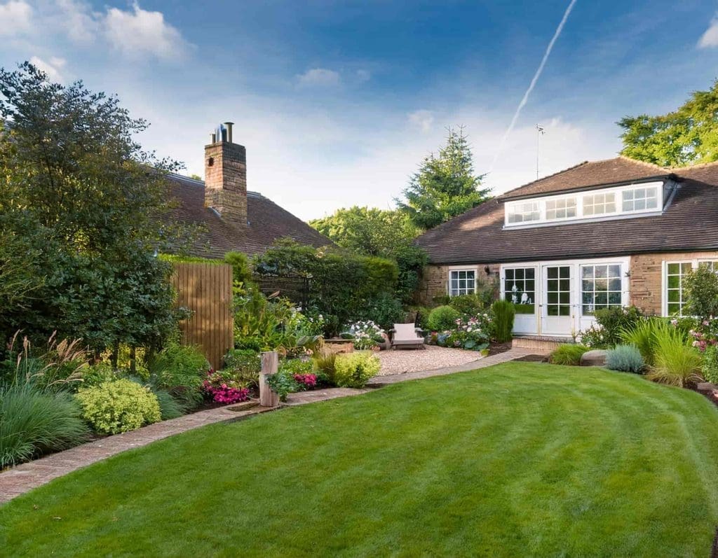 A well-maintained backyard garden with lush green grass, blooming flowers, shrubs, a paved path, and outdoor seating in front of a brick house with large windows under a blue sky.