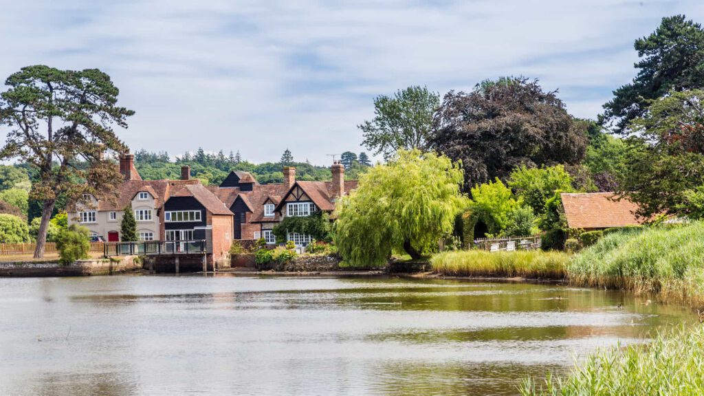 A tranquil village scene with charming houses and lush trees beside a calm river under a partly cloudy sky. Grassy banks and a red-roofed building are in the foreground.