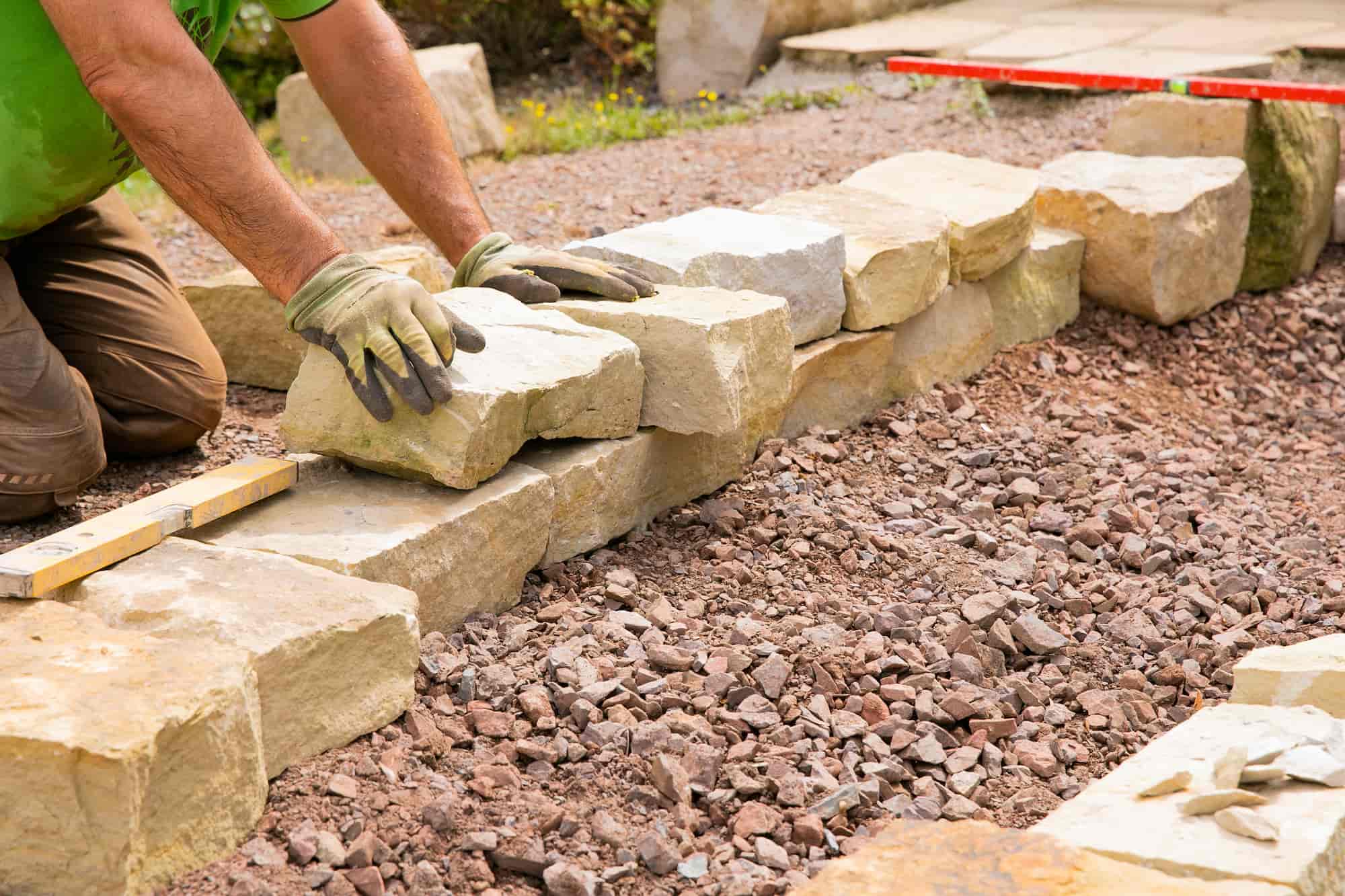 A person wearing gloves is kneeling on gravel, placing large stones in a row to build a stone wall. Measuring tools are visible on the ground beside the wall.