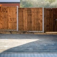 A wooden fence with a closed gate separates a paved area from a grassy yard. The fence is supported by concrete posts and stands beside a red brick building.