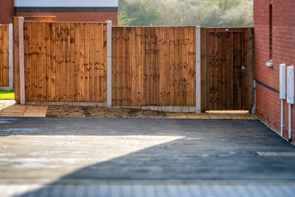 A wooden fence with a matching gate stands between two brick buildings, separating a paved area in the foreground from a grassy area in the background. Sunlight casts shadows on the ground.