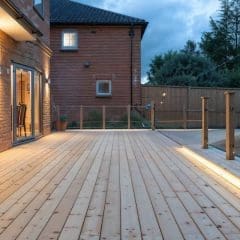 A spacious wooden deck with modern glass and wood railing, attached to a brick house. Warm outdoor lights illuminate the area at dusk, with a fenced backyard and trees in the background.